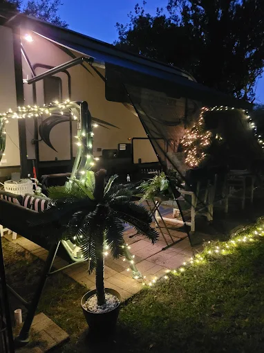 A cozy camper setup at night, decorated with string lights on the awning and a faux palm tree. Outdoor chairs and a table are arranged on a small patio, creating a warm, inviting atmosphere.