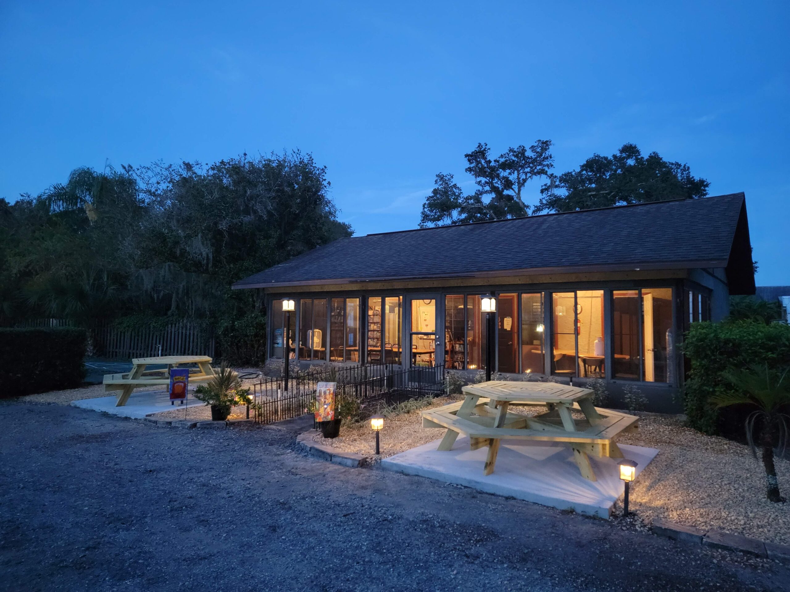 A cozy restaurant with large windows is lit warmly at dusk. Picnic tables sit outside on gravel, surrounded by small lights and trees, creating a welcoming atmosphere.